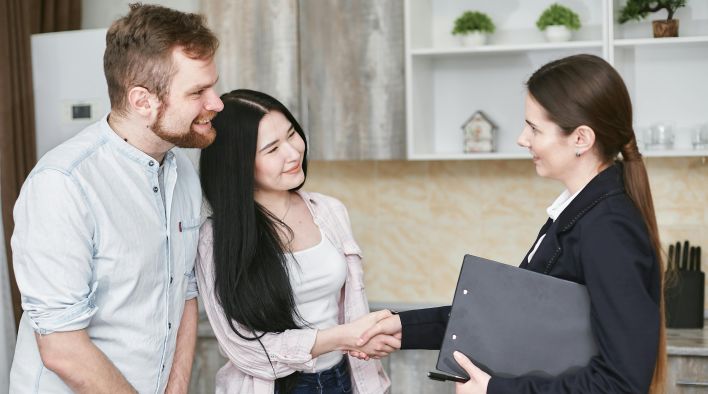 A real estate agent greets a couple with a handshake in a modern home interior.